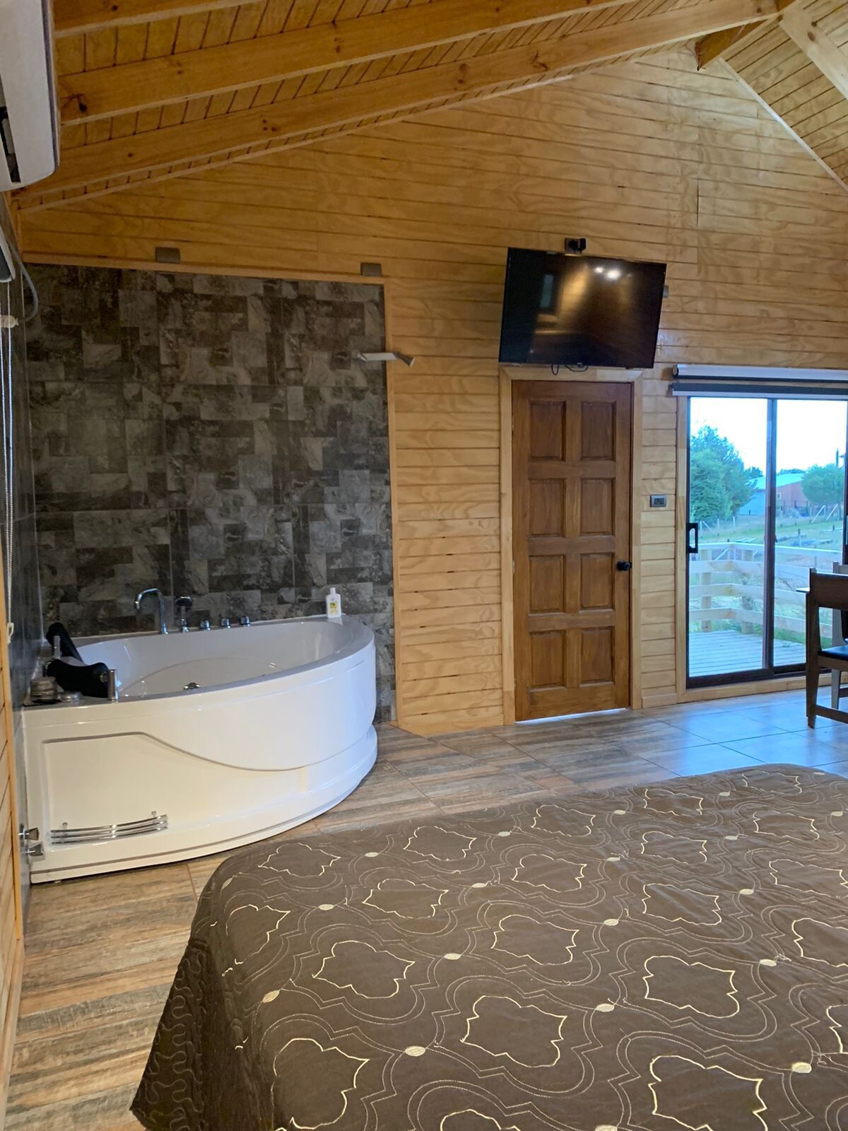 A spacious bedroom is equipped with a large bed and a flat-screen television mounted on the wall. A contemporary bathtub is positioned nearby, while sliding glass doors provide access to outdoor views. Natural wood accents create a warm atmosphere.