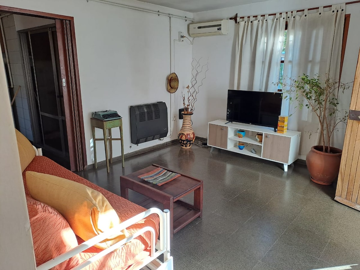 A living area features a comfortable orange sofa paired with a wooden coffee table. Natural light fills the space, highlighting a television unit with decorative elements. A plant adds a touch of greenery, and light curtains softly frame a window, enhancing the room's openness.