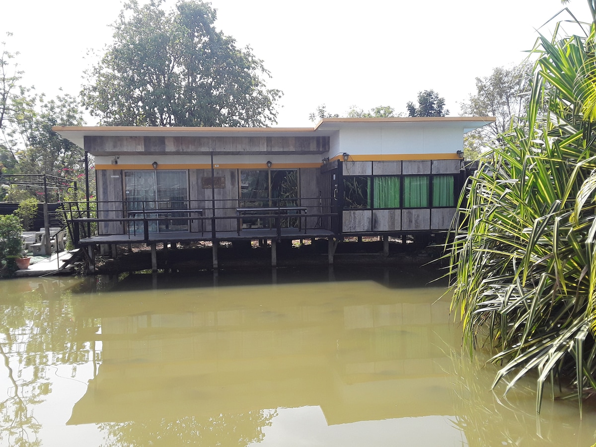 A tranquil waterside home is seen, featuring a wooden deck that extends over a calm body of water. The structure showcases large glass windows, allowing natural light to fill the interior. Lush greenery surrounds the property, enhancing the peaceful setting.
