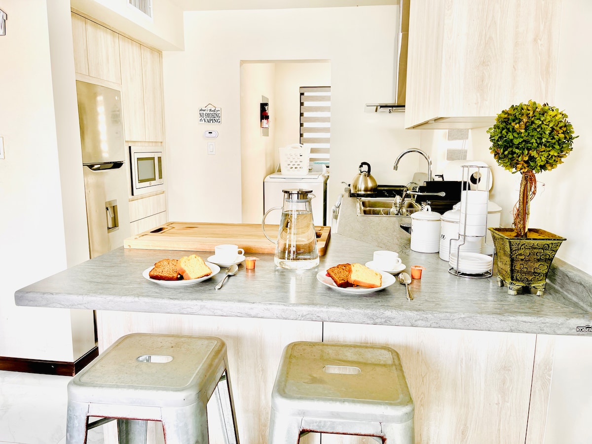 A modern kitchen features a marble countertop with two stools underneath. Plates with pastries and cups are set on a wooden cutting board. Stainless-steel appliances and kitchen utensils are organized, with a small potted plant adding a touch of greenery.