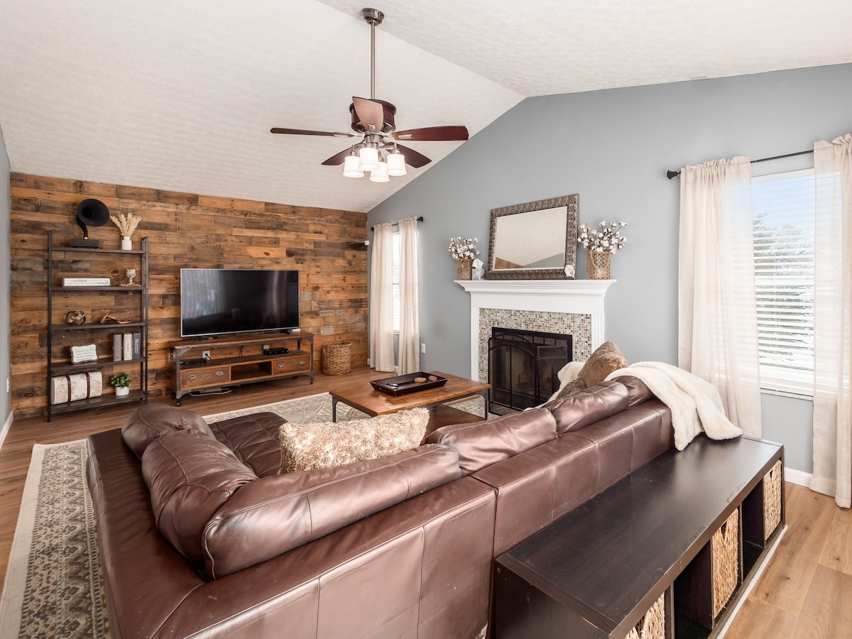 A spacious living area features a large, dark leather sectional sofa. A wooden shelf holds decorative items and a television is mounted on a rustic wood wall. Natural light fills the room through large windows, complemented by light curtains.