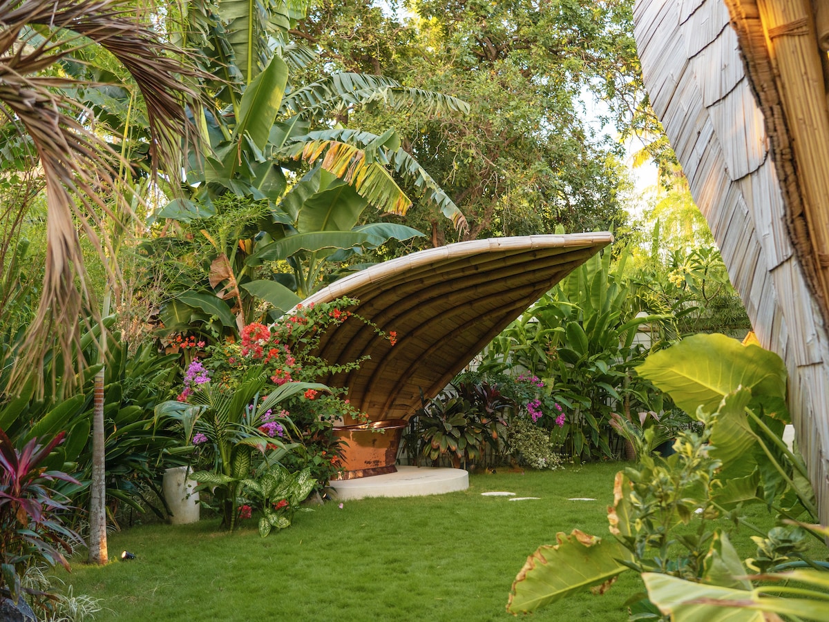A unique architectural feature of bamboo is set within a lush garden filled with tropical plants and vibrant flowers. The structure's curved roof is complemented by surrounding greenery, creating a serene and inviting outdoor space.