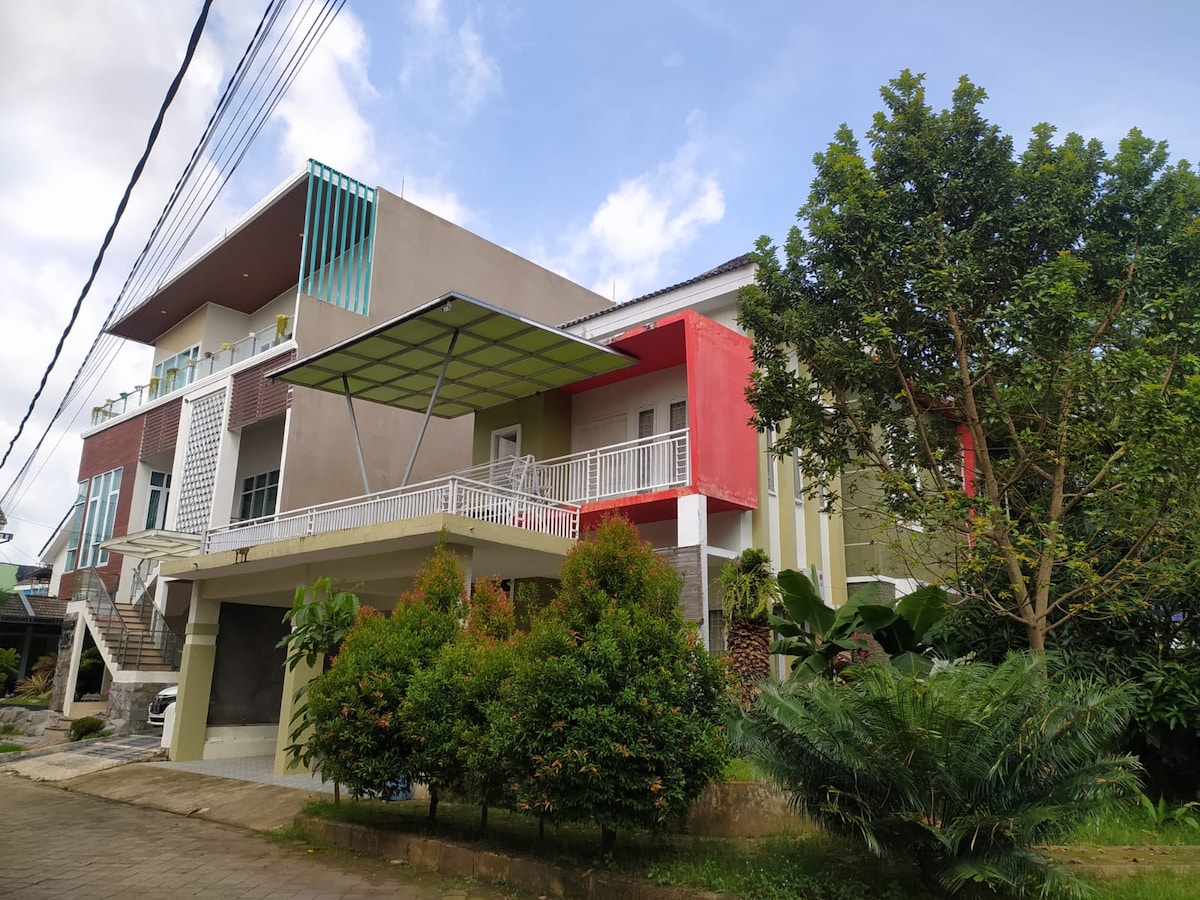 A modern two-story building features a mix of bold colors, including red and green accents. A spacious balcony with a unique canopy is visible above the entrance, surrounded by well-maintained greenery and shrubs, creating a serene courtyard atmosphere.