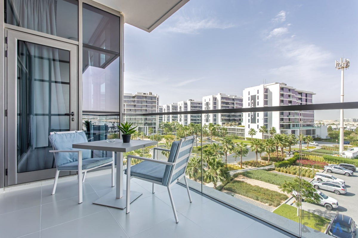 A balcony is shown featuring a small table and two light blue chairs. A potted plant adds a touch of greenery. The view includes landscaped gardens and residential buildings in the background, with a clear sky above enhancing the peaceful setting.