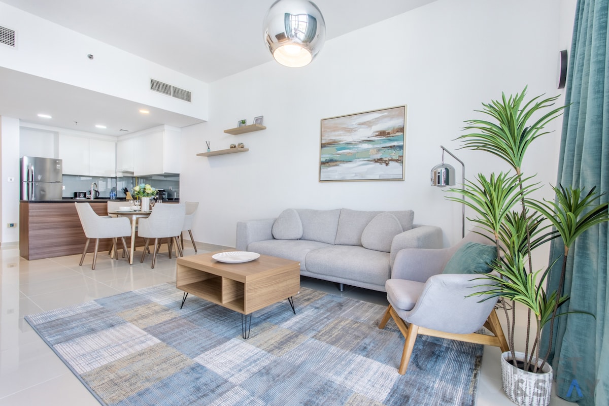 A modern living area features a light gray sofa and an armchair situated on a patterned area rug. A wooden coffee table is centrally placed, with a shelf above showcasing decor. A dining table for four is visible in the background, alongside a bright kitchen.