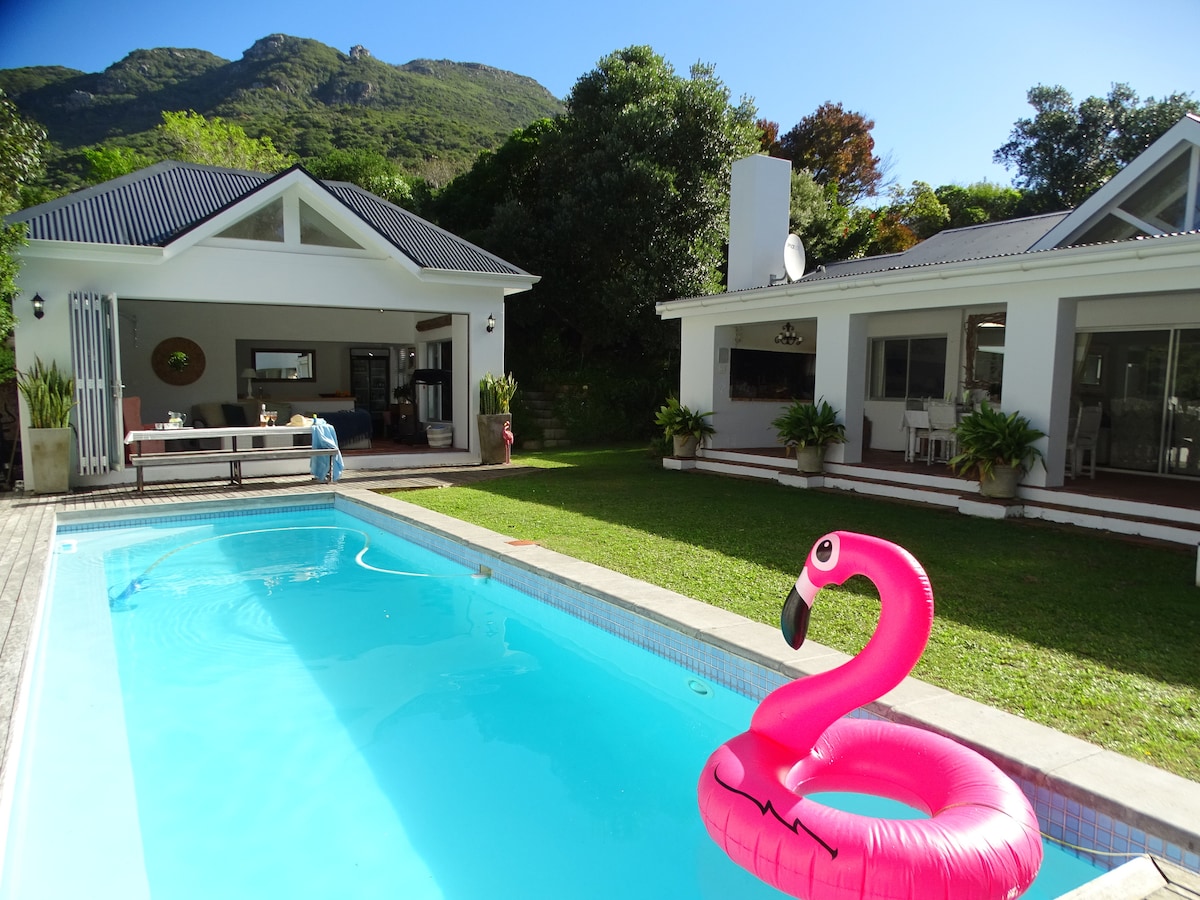 A large solar heated pool is presented, featuring a pink flamingo float in the clear water. Lush greenery surrounds the pool area, complemented by a spacious outdoor patio and modern lounge furniture. Two separate buildings are visible, showcasing a harmonious blend with the natural landscape.