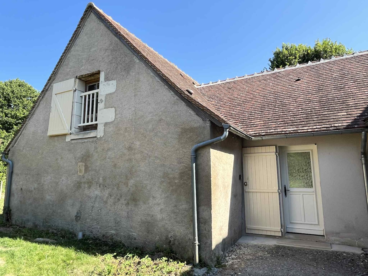 The exterior of the house is depicted, showcasing the rustic charm of the building with a sloping roof and a combination of textured stone and plaster walls. A spacious entrance door is visible, complemented by a window above it, framed by traditional shutters.