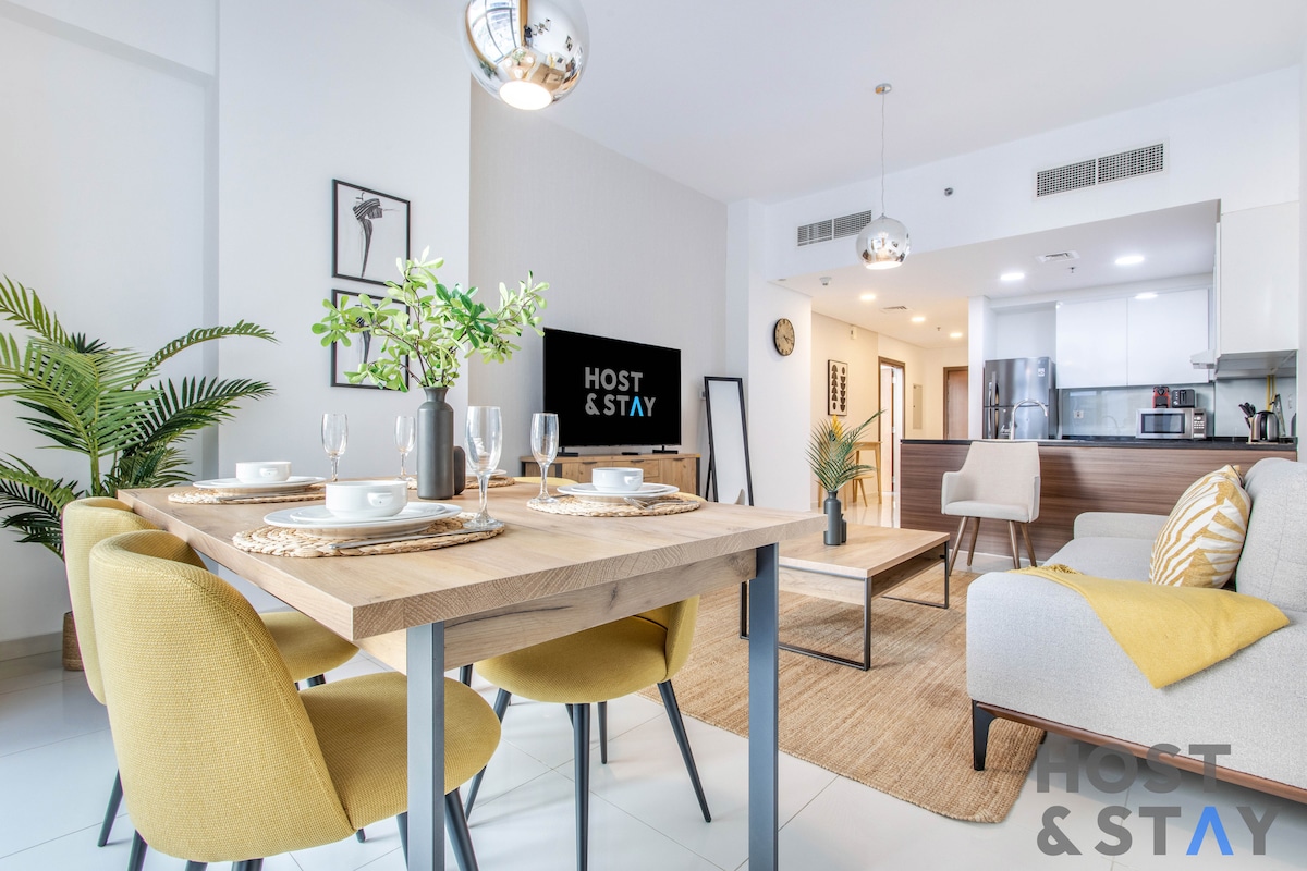 An inviting dining area is set with a wooden table and four yellow chairs, surrounded by potted greenery. A modern television is positioned on a neutral wall, while an open kitchen area is visible in the background, showcasing sleek appliances and cabinetry.