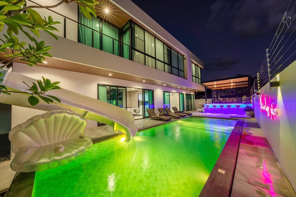 The private pool features a vibrant green hue illuminated by underwater lighting, complemented by a fun waterslide. Loungers are positioned along the pool's edge, with a stylish bar area in the background displaying colorful lighting against a modern villa façade.