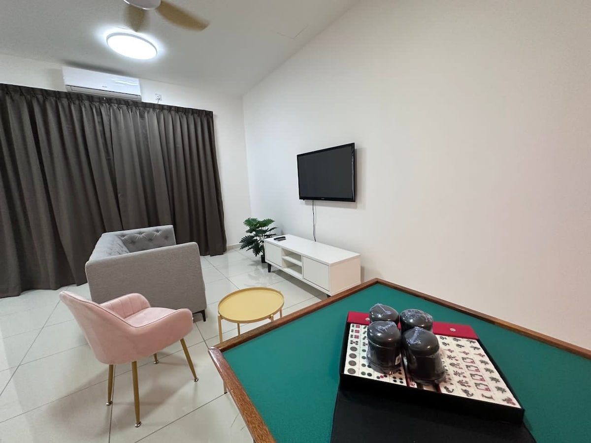 A simple lounge area features a gray sofa and a pink accent chair, accompanied by a small yellow side table. A wall-mounted television is positioned above a white media console. The space is illuminated by a ceiling light, with large dark curtains covering a window.