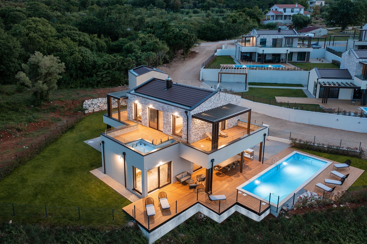 An aerial view of the two-story villa showcases a spacious layout with a swimming pool and wooden deck. Lush greenery surrounds the property, while sun loungers are positioned near the pool. The structure's modern design is highlighted by large glass doors and a striking stone exterior.
