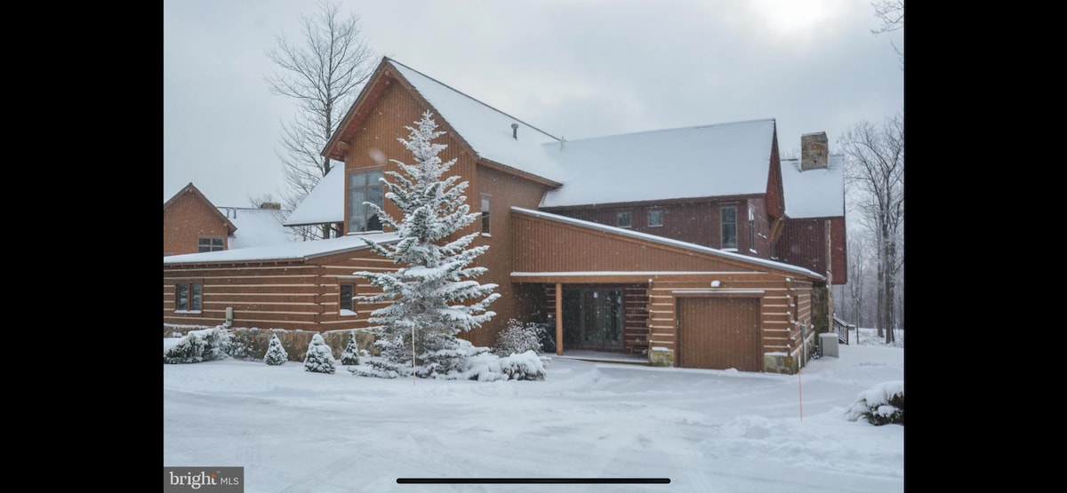 A spacious cabin is showcased amidst a snowy landscape, featuring a combination of timber and logs. The roof is blanketed in fresh snow, and a snow-covered evergreen tree stands prominently in front, enhancing the wintry scene. A garage is visible, with a plowed path leading to the entrance.