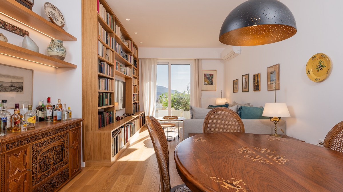 A well-appointed living area is highlighted by a spacious wooden dining table and stylish chairs. A bookshelf filled with various books lines one wall, while light pours in through glass doors leading to a balcony. Decorative items and art pieces add character to the space.