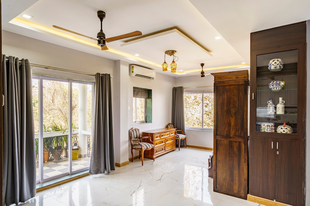 A bright and spacious living area features elegant marble flooring, with large windows allowing natural light to fill the room. Traditional furnishings, including a wooden seating area and decorative cabinet, complement the modern ceiling fixtures. Curtains frame the windows, enhancing the warm ambiance.