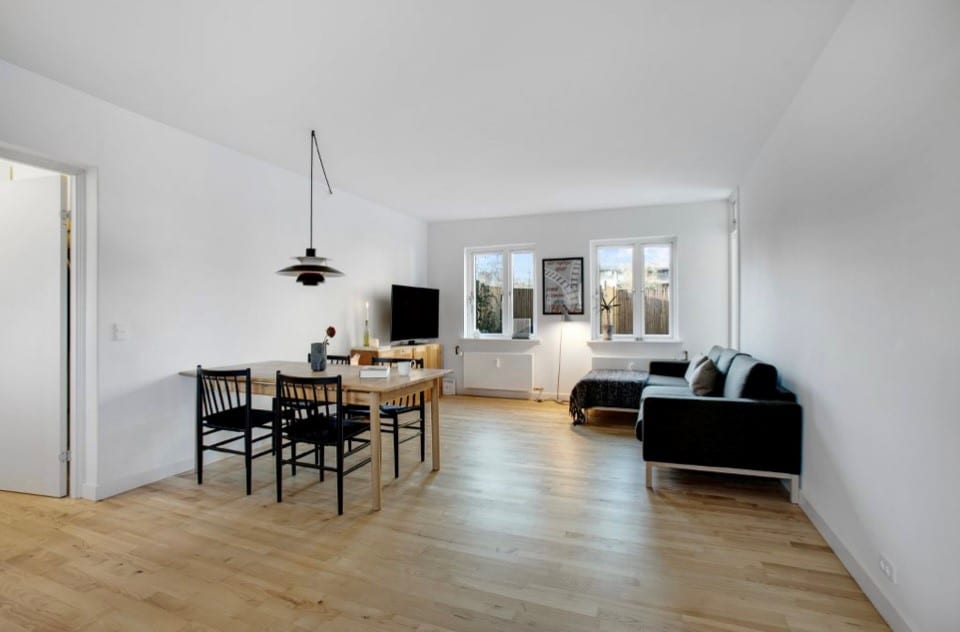 An open living space features wooden flooring, with a dining table and black chairs positioned centrally. A television is mounted on the wall, opposite a cozy sofa. Natural light enters through three large windows, providing a view of the outdoor greenery.