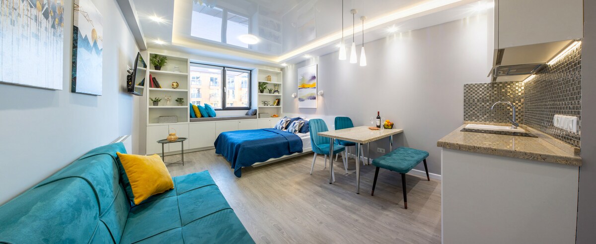 A modern studio features a spacious layout with a blue bedspread and teal seating. A dining area is equipped with a table and two chairs. Natural light enters through a large window, reflecting on sleek cabinetry and a compact kitchen area. Decorative items adorn the shelves.