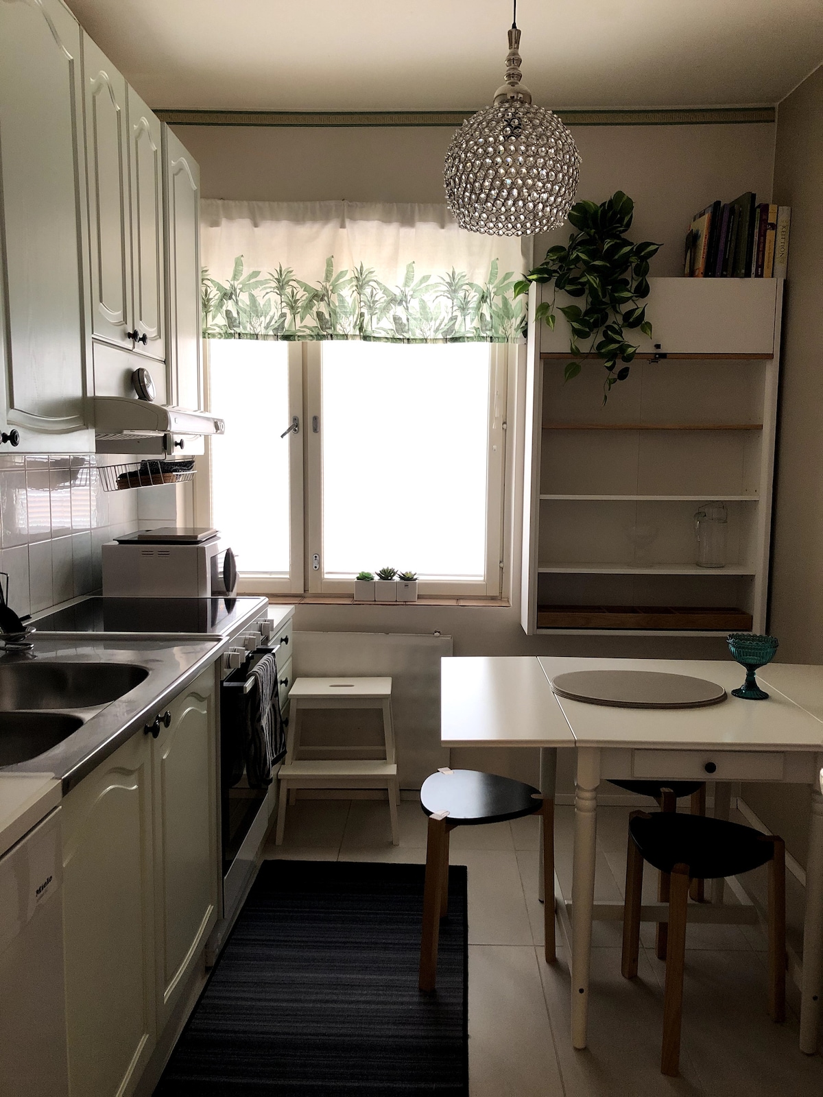 A compact kitchen is showcased, featuring white cabinetry and stainless steel appliances. Natural light enters through a window adorned with a light curtain. A small dining table with two chairs is positioned beside a tall bookshelf, complemented by decorative green plants.
