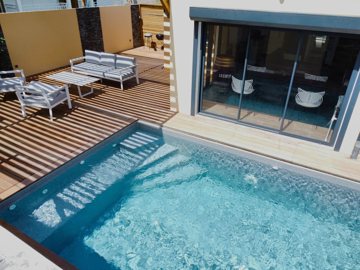A private pool features clear blue water, surrounded by wooden decking and inviting lounge seating. Sunlight creates patterns through the pergola overhead, enhancing the serene atmosphere of the outdoor space.