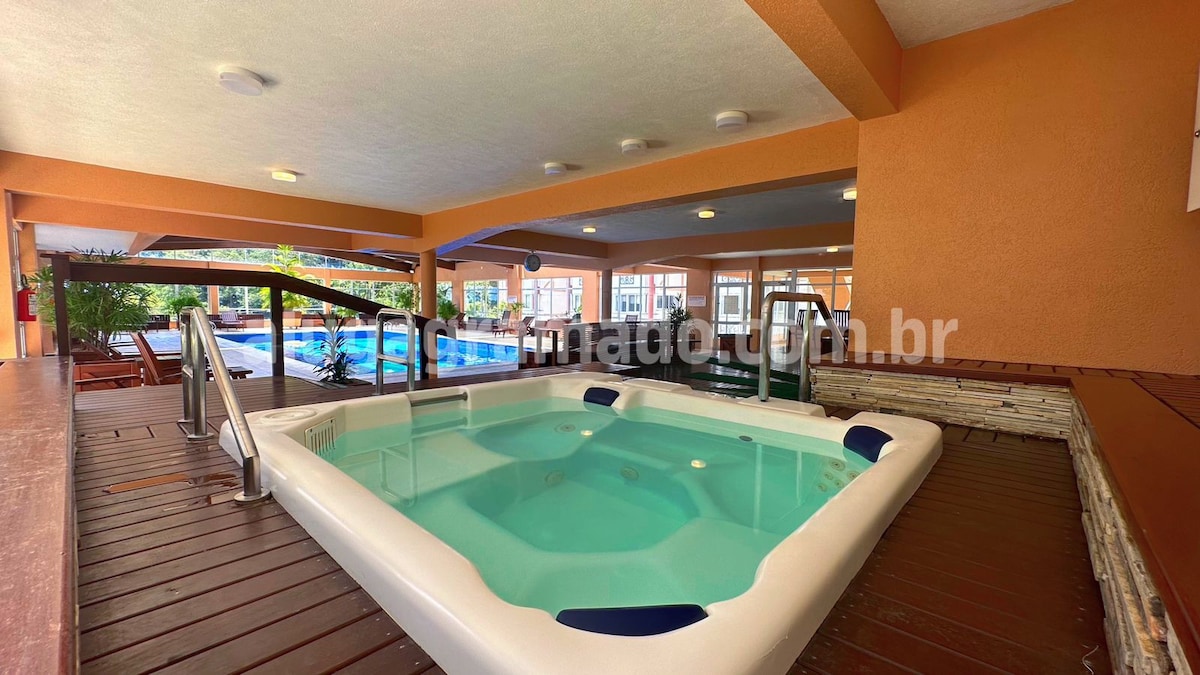 A serene indoor hot tub is set within a well-lit area, featuring a smooth surface and accessible handrails. Surrounding the hot tub, a wooden deck provides a warm contrast, while glimpses of outdoor pools are visible in the background.