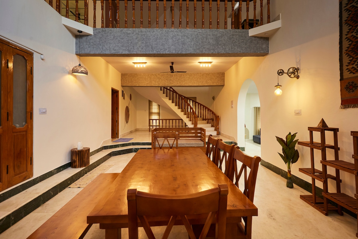 An open-plan dining area features a large wooden table surrounded by multiple chairs. Natural light brightens the space, highlighting the staircase in the background. Decorative elements include a small plant and a sleek wooden shelf, contributing to a serene and inviting environment.
