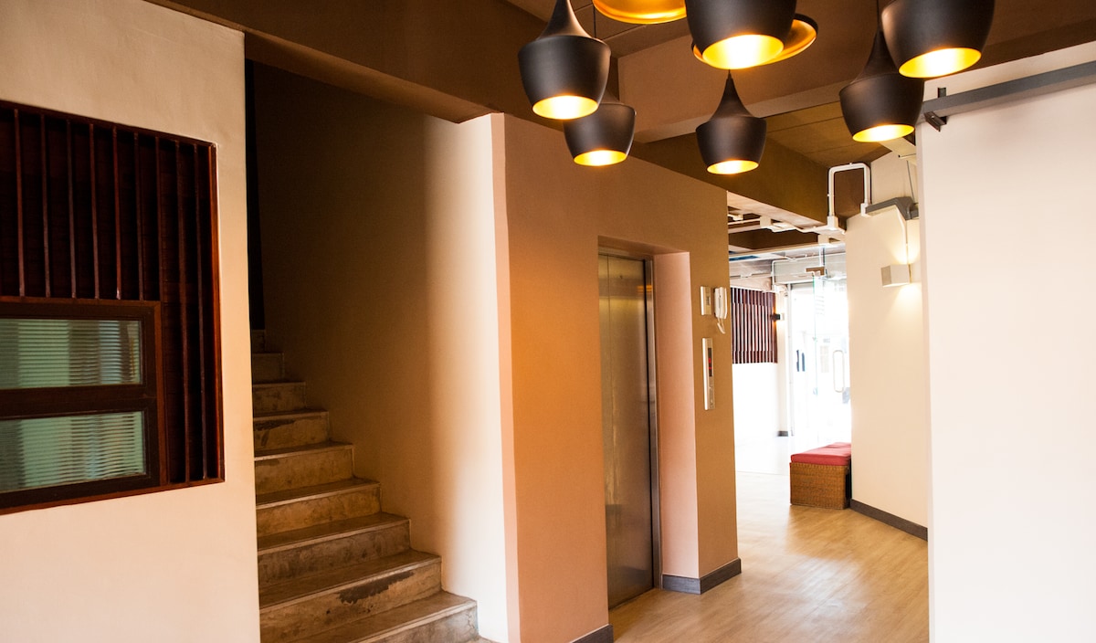 A well-lit corridor features a modern elevator and an adjacent staircase. The walls are painted in warm, neutral tones, and overhead, contemporary pendant lights create a welcoming ambiance. Natural light filters in from the open area at the end of the hallway.