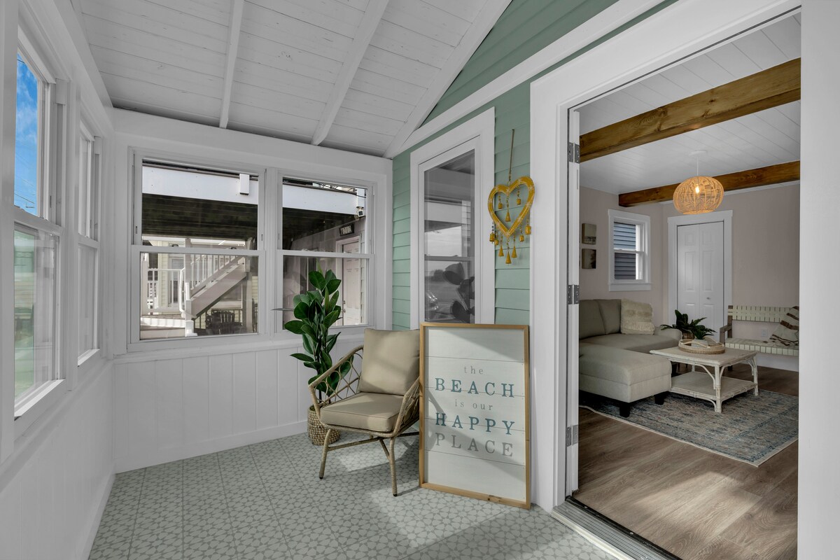 A cozy front porch features a light-colored chair and a decorative sign that reads 'the beach is our happy place.' Large windows allow natural light to fill the space, while an inviting view into the living area is visible through an open doorway.
