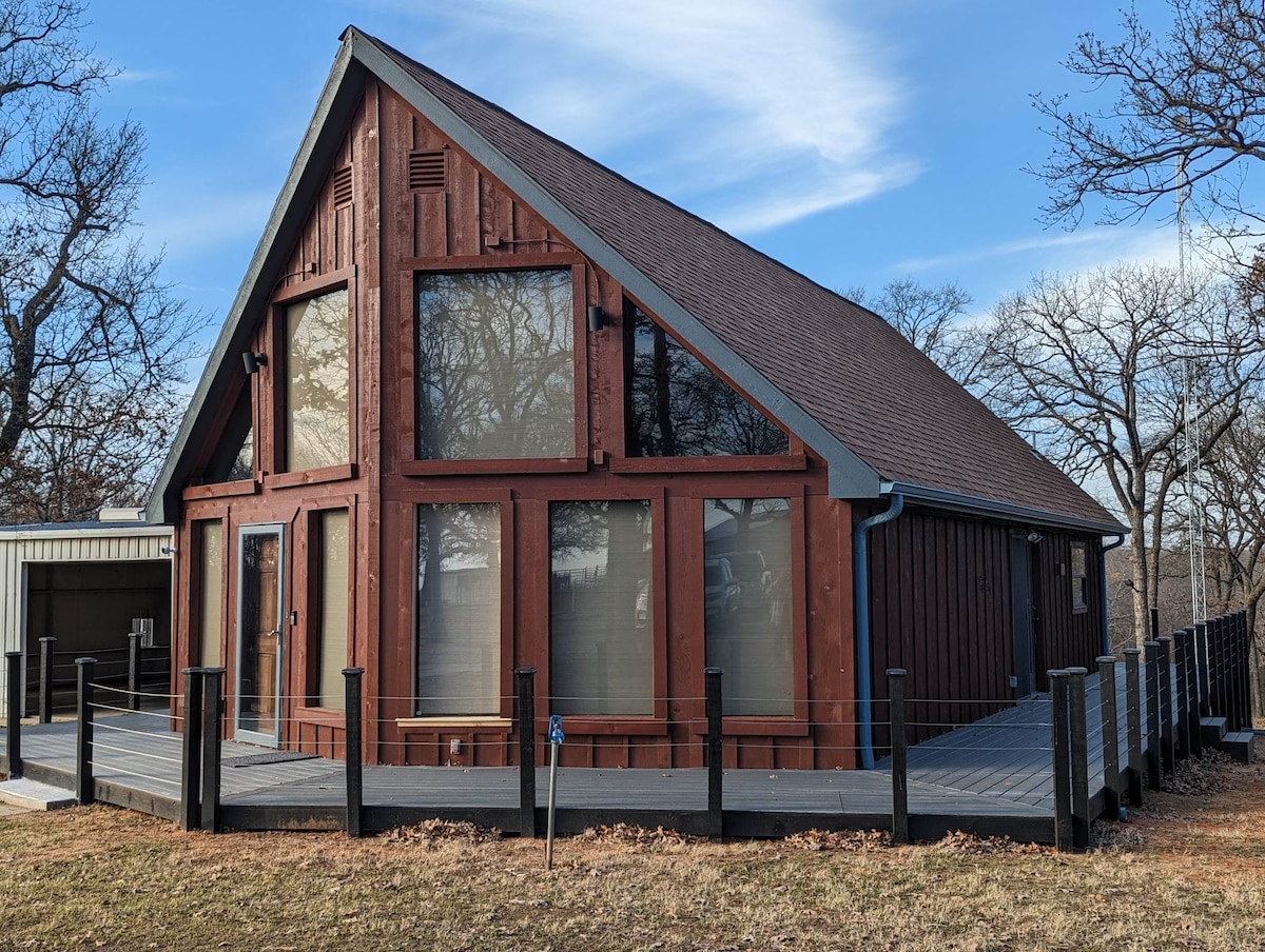 A-frame cottage features a striking red-brown exterior and large windows, providing ample natural light. The expansive deck is bordered by a simple black railing, and the surrounding landscape includes sparse trees and open space, suggesting a serene outdoor setting.
