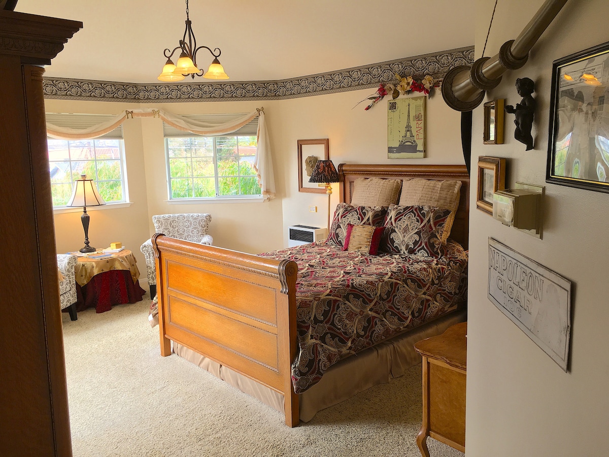 A spacious bedroom features a wooden sleigh bed with intricate patterned bedding. Natural light fills the room through two windows adorned with elegant drapery. A comfortable sitting area with a side table and lamp is positioned beside a cozy chair, creating an inviting nook.