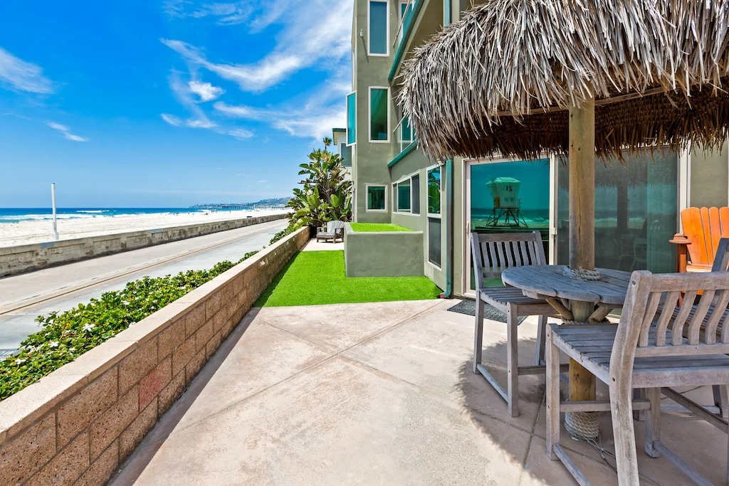 A spacious beachfront patio features a thatched-roof overhang, providing shade over a wooden table and chairs. Lush greenery and ocean views are visible in the background, with the waves gently lapping the shore.