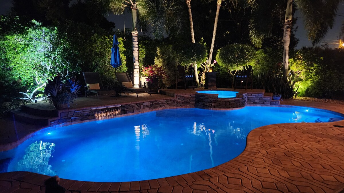 The outdoor area features a heated saltwater pool with a waterfall, illuminated in soft blue tones. A hot tub is situated nearby, surrounded by lush landscaping and palm trees. Subtle lighting highlights the palm fronds, creating a serene nighttime atmosphere.