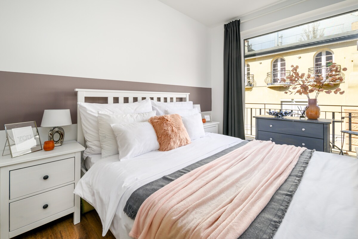 A bright bedroom is furnished with a spacious bed adorned with pillows and a soft throw. Nightstands with lamps are positioned on either side of the bed. A large window offers views of an exterior balcony, complemented by light curtains framing the scene.