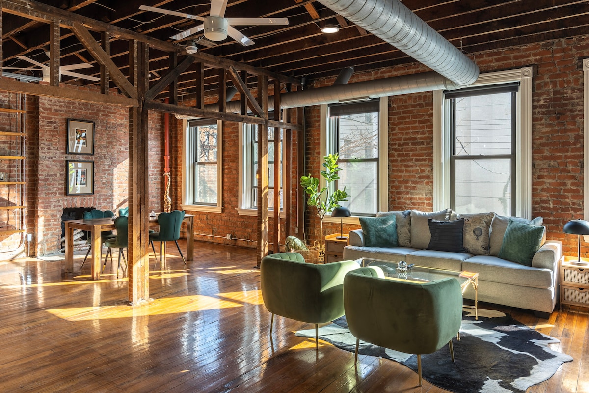 An open loft space is showcased with exposed brick walls and wooden beams, featuring large windows that allow natural light to fill the room. A stylish sectional sofa and modern armchairs surround a glass coffee table, while a dining area with patterned chairs is visible in the background.
