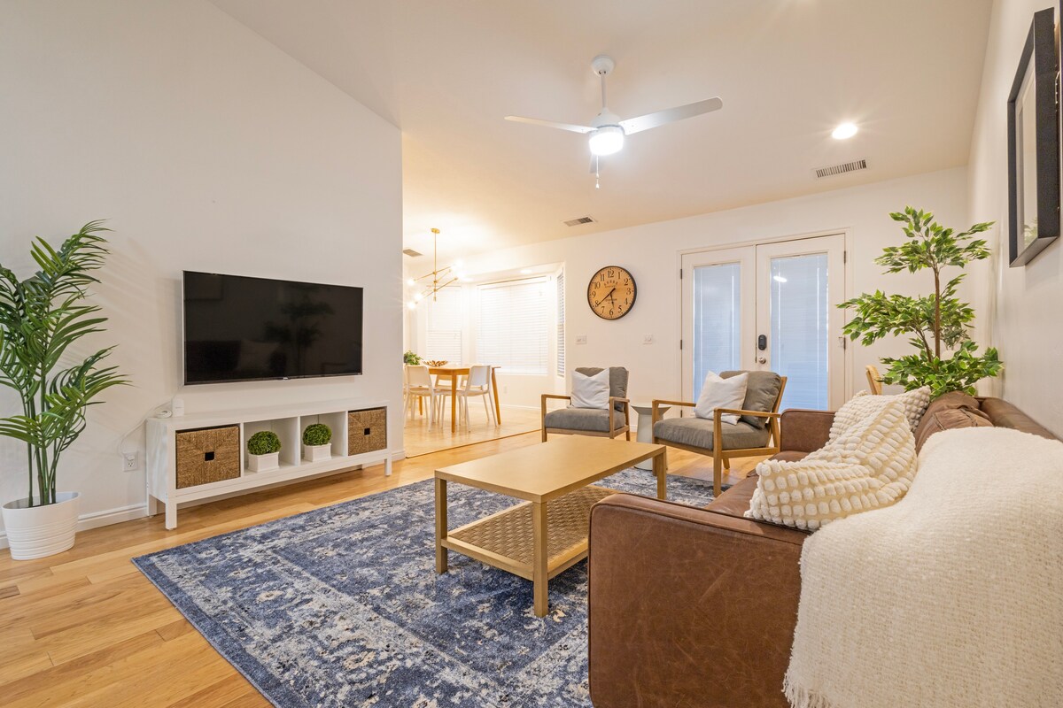 A welcoming living area features a brown sofa with a light blanket draped across it. A large flat-screen television is mounted on a white media console, and two armchairs sit beside a wooden coffee table. Natural light enters through French doors, illuminating the space.