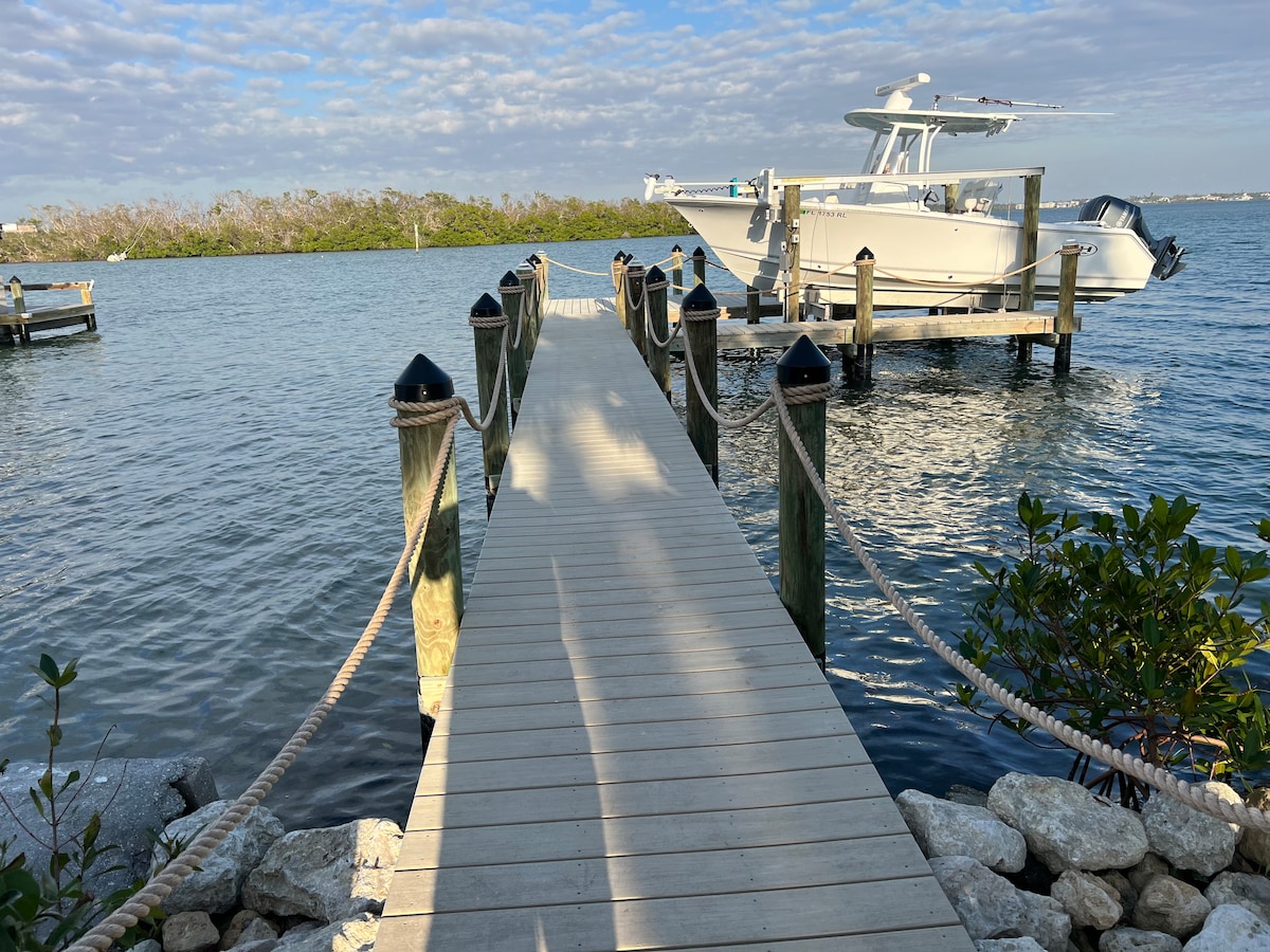A wooden dock extends over calm waters, leading to a boat secured at the end. The surroundings include gentle waves and patches of greenery. Light clouds create a textured sky above, adding a serene backdrop to the scene.