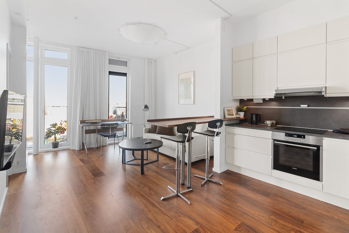A modern kitchen and living area showcase sleek cabinetry and a spacious countertop, complemented by elegant wooden flooring. A dining space with a round table and chairs is positioned near large windows, allowing natural light to illuminate the room.
