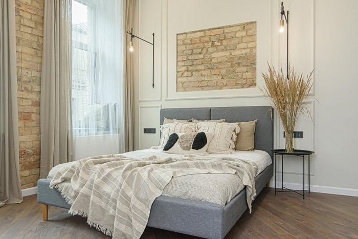 A spacious sleeping area features a comfortable bed dressed in soft linens and decorative pillows. Exposed brick wall adds character, while large windows allow natural light to illuminate the room. A minimalist bedside table holds a decorative plant, enhancing the contemporary design.