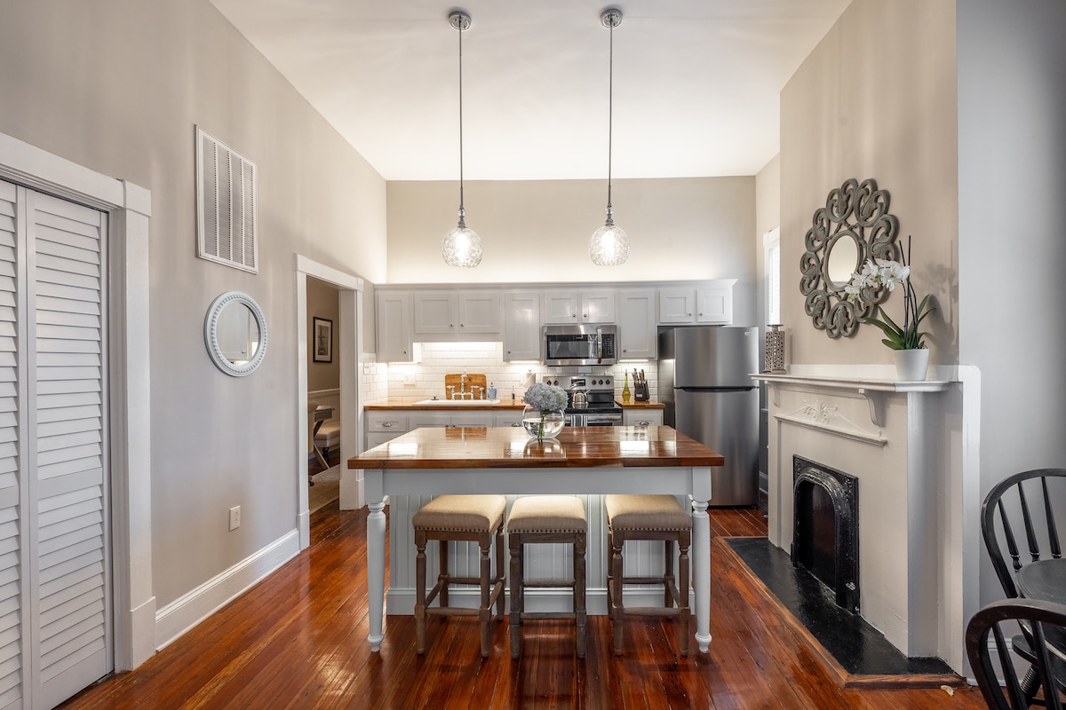 A spacious kitchen is highlighted by a central wooden dining table surrounded by four woven stools. Pendant lights hang above, illuminating the area. Modern appliances, including a stainless-steel refrigerator and oven, complement the white cabinetry. Warm hardwood flooring adds a welcoming touch.