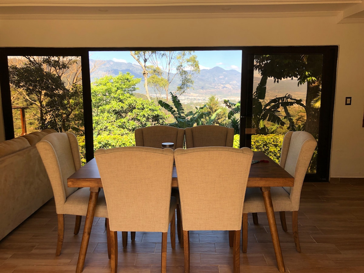 A wooden dining table is surrounded by six upholstered chairs, set against large glass doors that offer views of the lush greenery and distant mountains. Natural light fills the space, creating a bright atmosphere.