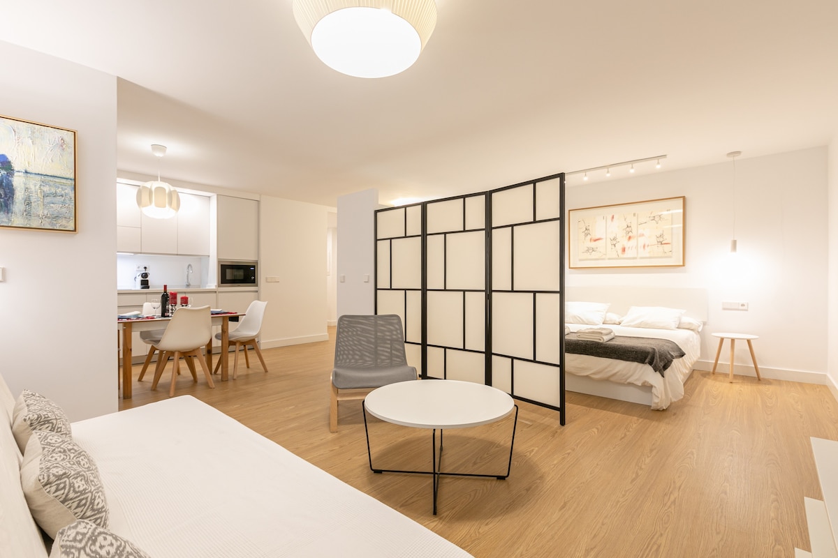 An open living space showcases a modern design, featuring a comfortable sofa and a sleek coffee table. A room divider separates the sleeping area, furnished with a spacious bed neatly made, while a dining table and chairs are visible in the background.