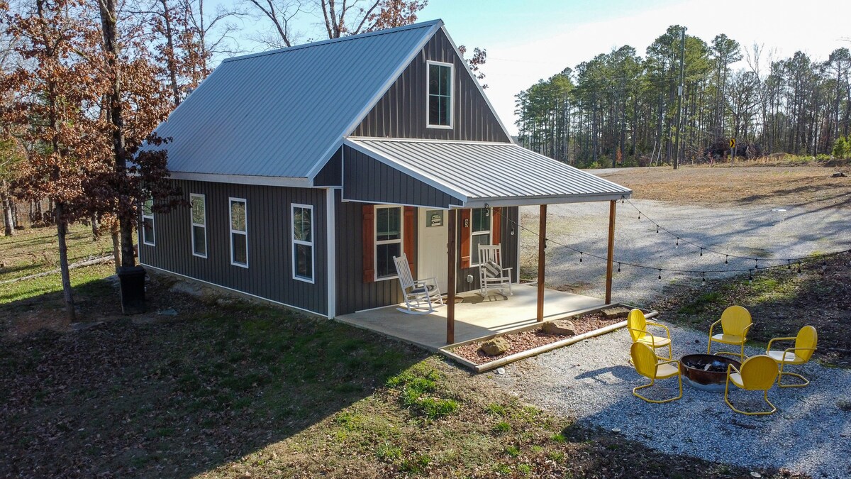 This charming cottage showcases a modern exterior with a metal roof and dark wooden siding. A spacious front porch is adorned with two rocking chairs, inviting relaxation. A fire pit surrounded by bright yellow chairs sits in the outdoor area, framed by a natural landscape of trees.