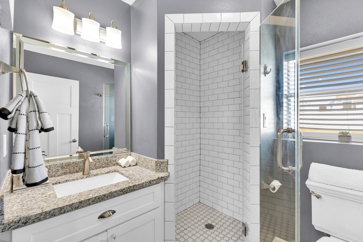 A modern bathroom is equipped with a spacious shower featuring white subway tiles and a sleek glass enclosure. A granite countertop with dual sinks is complemented by bright light fixtures overhead. A towel hangs neatly on a rack, and a small potted plant adds a touch of greenery.