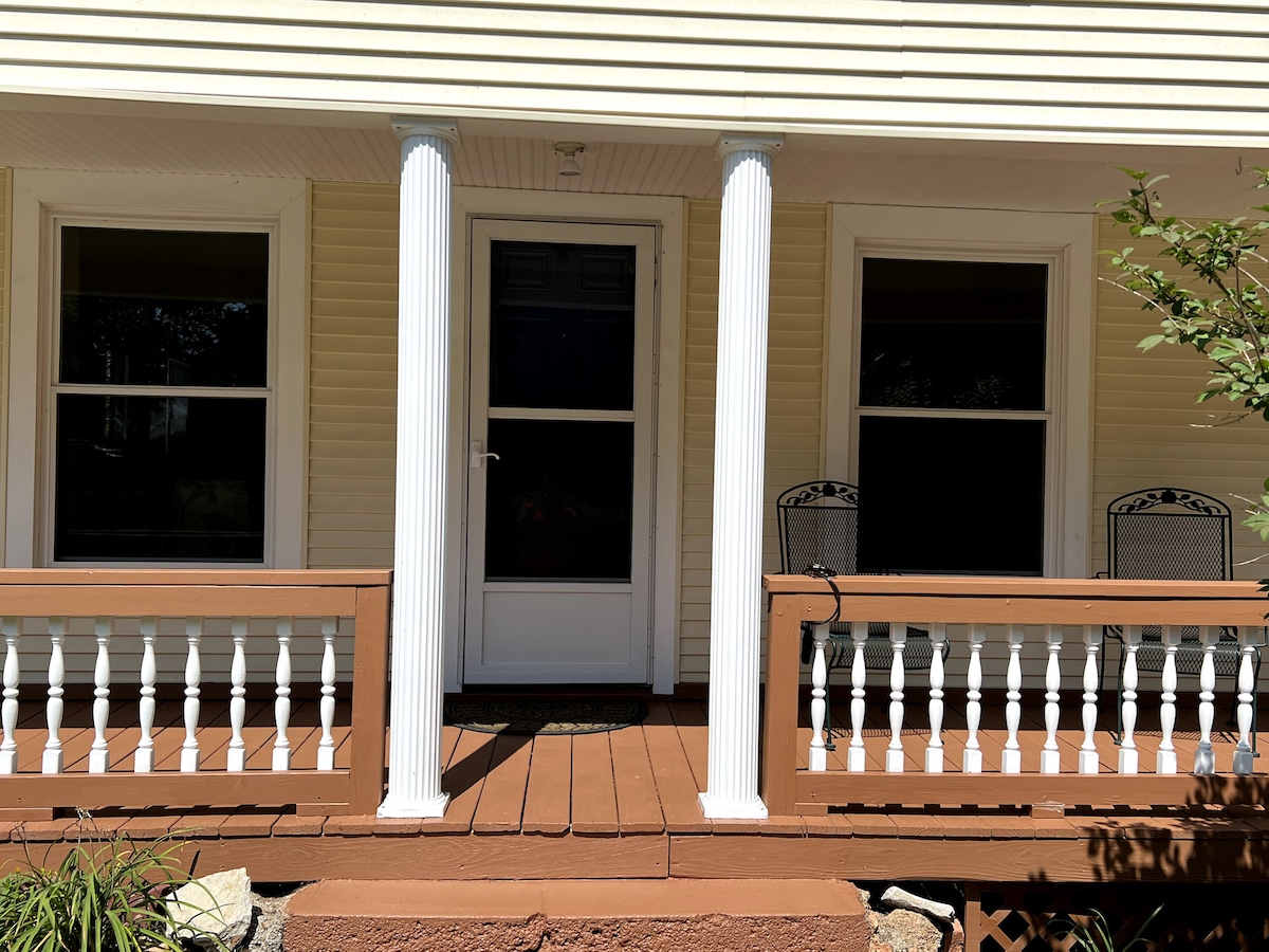 The cottage entrance is framed by two white columns, leading to a front door. Two black wrought-iron chairs are placed on the porch, alongside a wooden railing featuring a row of spindles. Large windows on either side allow natural light to illuminate the space.