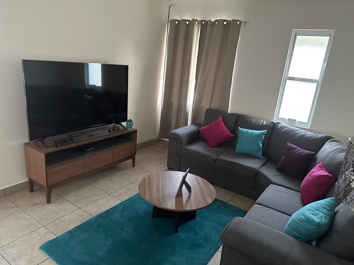 A modern living area features a comfortable sectional sofa adorned with colorful pillows. A sleek coffee table sits at the center on a soft area rug. A large television is positioned on a wooden console, with natural light streaming through a nearby window.