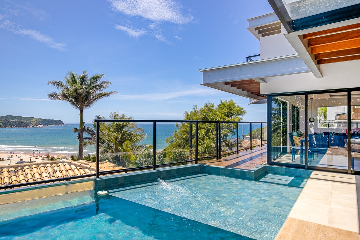 An inviting pool area features a sleek infinity edge, overlooking a sandy beach and clear blue ocean. Lush palm trees frame the view, adding a tropical touch. Large glass doors lead into the indoor living space, creating a seamless connection with the outdoors.