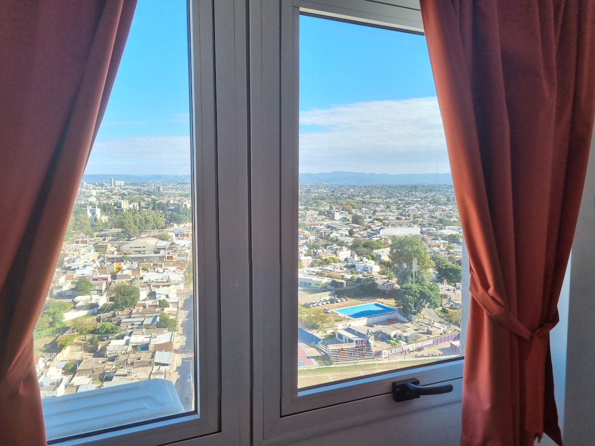A large window offers a panoramic view of the cityscape, showcasing a mix of residential areas and green spaces. Bright blue skies contrast with the colorful buildings and rooftops below. Soft, flowing curtains frame the window, complementing the view.