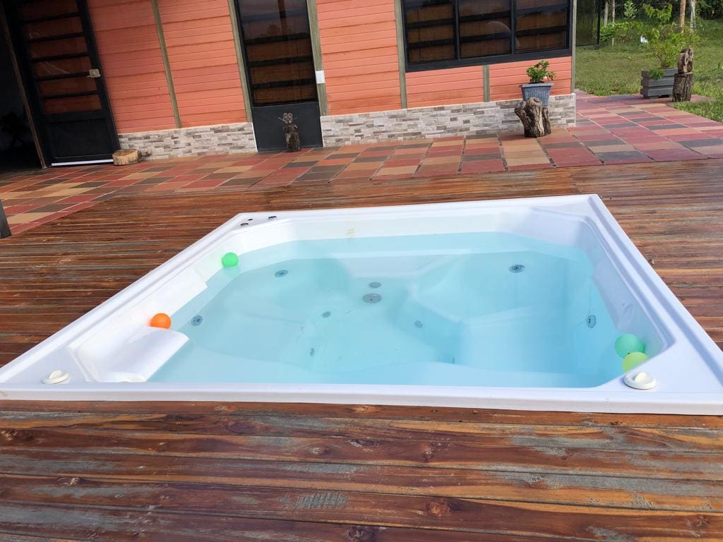 A white jacuzzi is positioned on a wooden deck, surrounded by nature. The water is clear, with colorful floating balls visible. The vibrant flooring consists of various tiles, and large glass doors lead to the interior space, enhancing the overall inviting atmosphere.