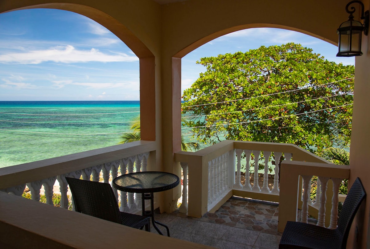 A private patio features a small round table accompanied by two chairs. Arched openings provide a view of the clear turquoise water and lush greenery, creating a serene seaside setting.