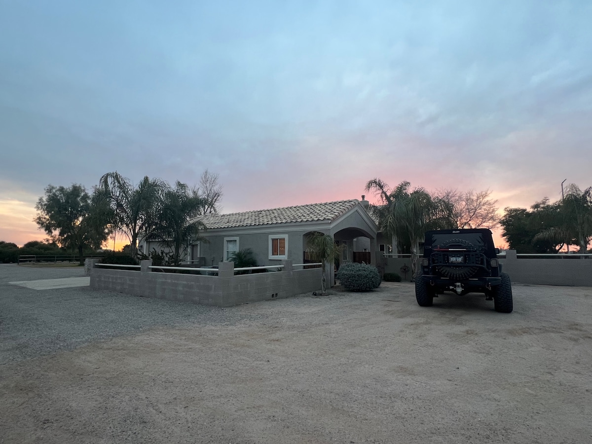 A serene evening scene showcases the casita set against a colorful sky at dusk. Tropical palm trees line the property, with a spacious gravel driveway leading to the entrance. A large vehicle is parked in the foreground, emphasizing the ample parking available.