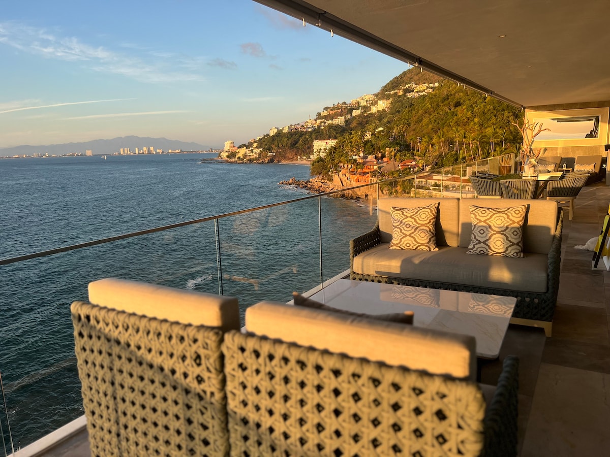 A spacious terrace offers stunning views of the ocean and coastline. Comfortable outdoor seating is arranged around a light-colored table, creating an inviting area for relaxation. The warm evening light casts a gentle glow on the scene, enhancing the tranquil coastal atmosphere.