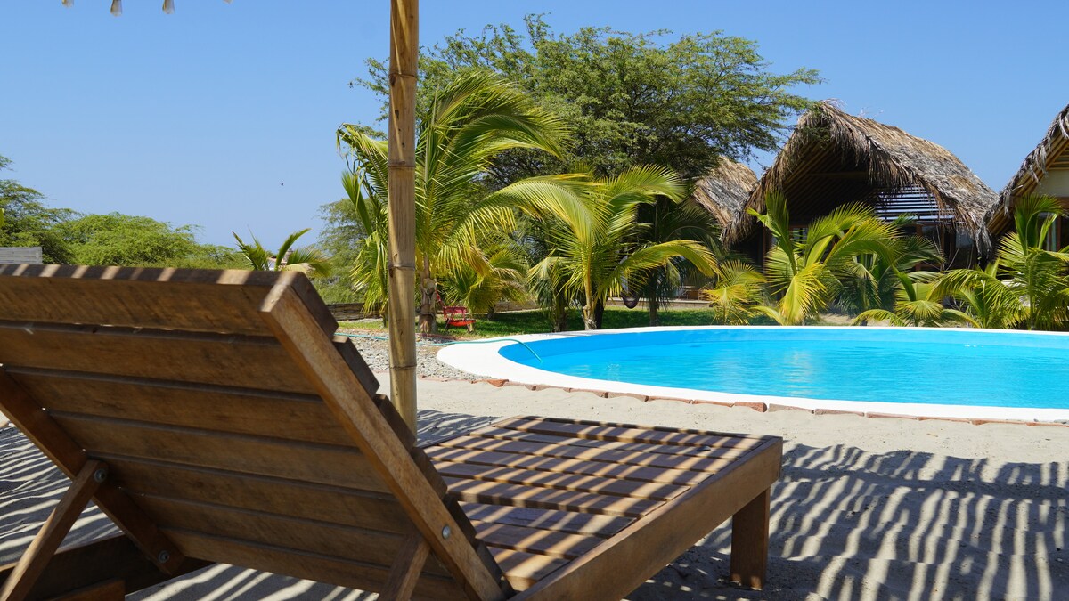 A wooden lounge chair is positioned beside a sparkling blue pool, with shadows cast by palm leaves overhead. In the background, thatched-roof bungalows and lush greenery can be seen, contributing to a serene outdoor setting.